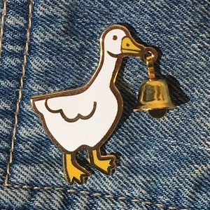 May include: A white goose enamel pin with yellow accents, holding a gold bell in its beak. The pin is on a blue denim background.