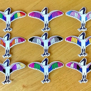 May include: Nine colourful stickers of a white bird with a black head and a long beak. The bird has wings with different coloured stripes, including rainbow, pink, grey, and blue.