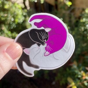 May include: A sticker featuring three cats in a yin yang design. The cats are black, white, and purple. The black cat is on the left, the white cat is on the right, and the purple cat is in the middle.
