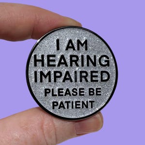 May include: A circular pin with a silver glitter surface and a black border. The pin features the text "I AM HEARING IMPAIRED PLEASE BE PATIENT" in black, bold lettering. The pin is held up against a purple background.