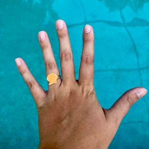 Puede incluir: Un anillo de oro con un diseño de concha. El anillo está en el dedo de una persona.