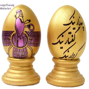 May include: Two golden eggs on stands, one decorated with a pink and purple image of a winged figure, the other with black calligraphy in Farsi.