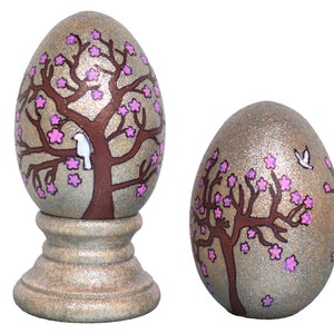 May include: Two decorative eggs with a glitter finish. Each egg features a brown tree with pink blossoms and a white bird. The larger egg is on a stand.