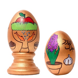 May include: Two golden eggs decorated with colorful paint. One egg features a red apple, green grass, and two orange fish. The other egg has a purple flower, green leaves, and a brown pot. Both eggs are on a white background.
