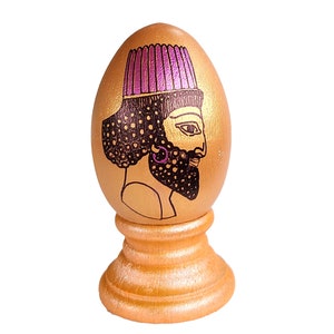 May include: A decorative golden egg with a painted illustration of a man's profile, featuring a purple headdress and black beard. The egg is displayed on a matching golden stand, creating a unique decorative piece.