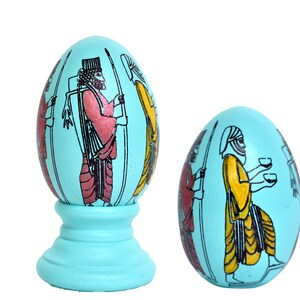 May include: Two decorative eggs with a turquoise blue background. The eggs are painted with black outlines and colorful figures of people in traditional clothing. One egg is on a turquoise blue stand.