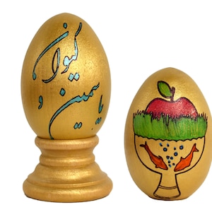 May include: Two golden eggs decorated with traditional Persian designs. One egg features a calligraphic inscription in blue and black, while the other depicts a red apple, green grass, and two orange fish in a bowl.