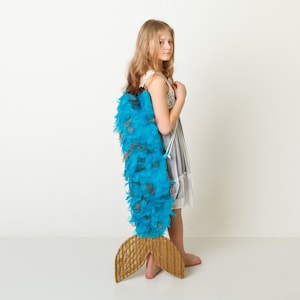 Puede incluir: Una niña pequeña con un vestido blanco y una cola de sirena de plumas azules y marrones.