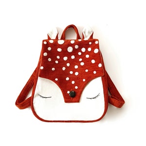 Puede incluir: Una pequeña mochila con forma de ciervo naranja y blanco con una cara blanca y pestañas negras. La mochila tiene un botón marrón en la nariz.