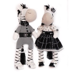 May include: Two black and white striped zebra stuffed animals. The zebra on the left is wearing a black and white striped shirt, a black belt, and black and white striped pants. The zebra on the right is wearing a black and white dress with a black and white plaid skirt.