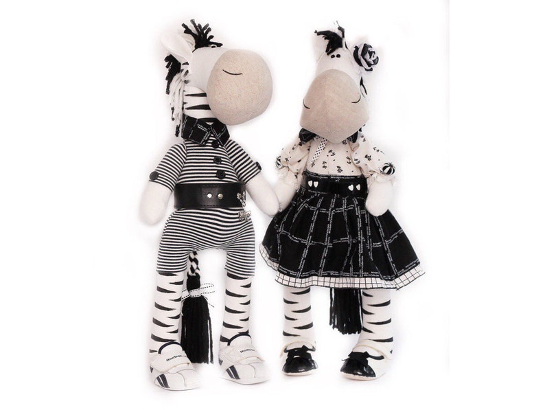 May include: Two black and white striped zebra stuffed animals. The zebra on the left is wearing a black and white striped shirt, a black belt, and black and white striped pants. The zebra on the right is wearing a black and white dress with a black and white plaid skirt.