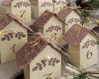 May include: A collection of small, cream-coloured house-shaped ornaments with brown corduroy roofs. Each house has a gold number and a decorative leaf design. A pearl is centered on each house. Gold string lights and a gold bow are attached to each house.