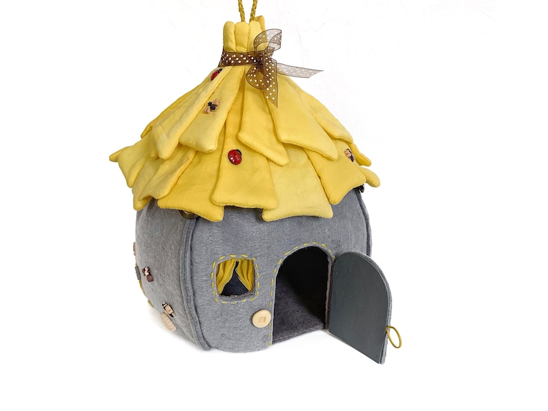 May include: A gray felt house with a yellow felt roof and a small door. The roof has a brown ribbon and a white polka dot bow. The house has a small window with a blue curtain.