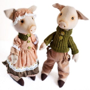 May include: Two handmade fabric pig dolls. The female pig doll is wearing a pink dress with white lace trim and a green collar. The male pig doll is wearing a green sweater and brown pants.