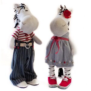 May include: Two plush zebra toys, one wearing a red and white striped shirt, blue jeans, and a chain, and the other wearing a blue and white striped dress with a red bow and a red flower.