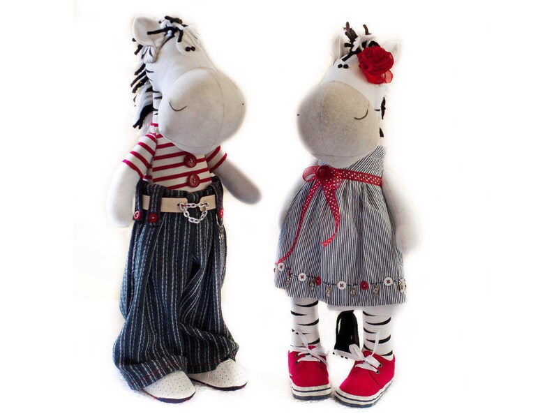 May include: Two plush zebra toys, one wearing a red and white striped shirt, blue jeans, and a chain, and the other wearing a blue and white striped dress with a red bow and a red flower.