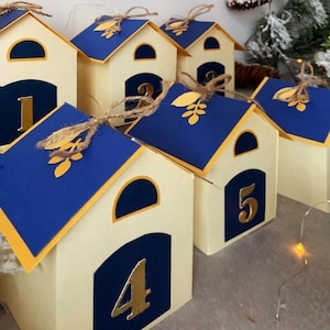 May include: Advent calendar featuring cream-coloured house-shaped boxes. Each house has a blue roof with a gold leaf design and a gold number. The boxes are decorated with a gold number from 1 to 5. The houses are tied with twine.