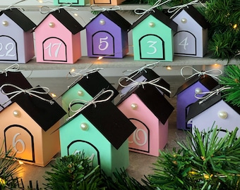 May include: Colorful house-shaped boxes with black roofs, each displaying a number from 1 to 22. The boxes are in pastel colors like pink, purple, and green, adorned with a pearl and a silver bow. Festive and decorative.