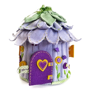 Könnte beinhalten: Ein lilafarbenes Filz-Feenhaus mit einem grünen Filzdach, einer lilafarbenen Filztür mit einem gelben Herzen und gelben und lilafarbenen Filzblumen.