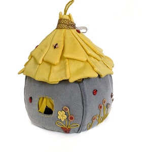 May include: A gray felt house with a yellow felt roof and embroidered flowers. The house has a small window and a red ladybug on the side.