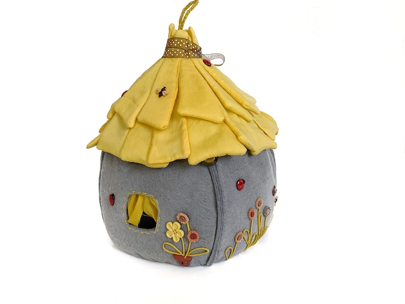 May include: A gray felt house with a yellow felt roof and embroidered flowers. The house has a small window and a red ladybug on the side.