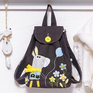 May include: A small grey canvas backpack with a flap closure and a yellow button. The backpack features an embroidered grey donkey holding a blue balloon and surrounded by yellow, white and green flowers.