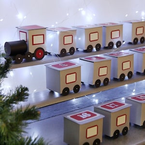 Puede incluir: Un calendario de adviento artesanal con forma de tren. La locomotora es negra y los vagones son grises con detalles rojos y pequeñas ruedas negras. Cada vagón está numerado. El tren está sobre una estantería de madera, con luces de hadas en el fondo.