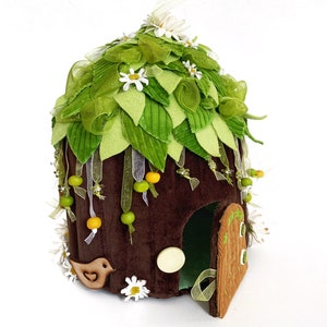 K&ouml;nnte beinhalten: Ein braunes Filz-Feenhaus mit einem gr&uuml;nen Filzdach, verziert mit wei&szlig;en Bl&uuml;ten, gr&uuml;nen Bl&auml;ttern und gelben Perlen. Das Haus hat eine kleine T&uuml;r und eine Vogelfigur.