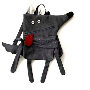 May include: A grey wolf-shaped backpack with white ears, black eyes, and a red pocket. The backpack has a zipper and a patterned fabric detail.
