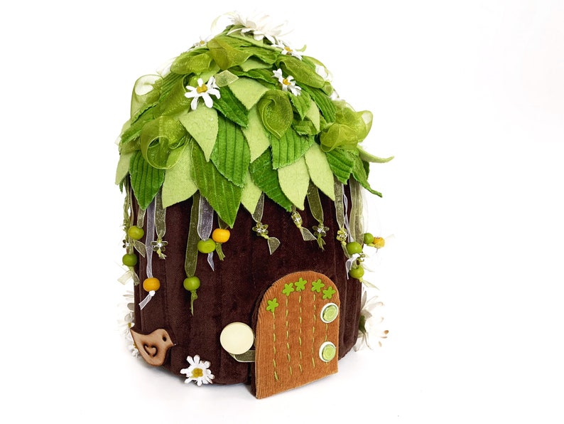 K&ouml;nnte beinhalten: Ein braunes Filz-Feenhaus mit einem gr&uuml;nen Filzdach, verziert mit gr&uuml;nen Bl&auml;ttern, wei&szlig;en Bl&uuml;ten und gelben und gr&uuml;nen Perlen. Das Haus hat eine kleine braune T&uuml;r mit gr&uuml;nem Filzrand und einem wei&szlig;en Filzkreis.