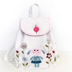 May include: White linen backpack with a pink button closure and embroidered flowers and a bunny with blue and pink accents.