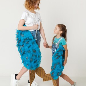 May include: Two girls are holding blue and brown fish-shaped props. The girl on the left is wearing a white t-shirt and denim shorts. The girl on the right is wearing a blue and white striped shirt and denim overalls.