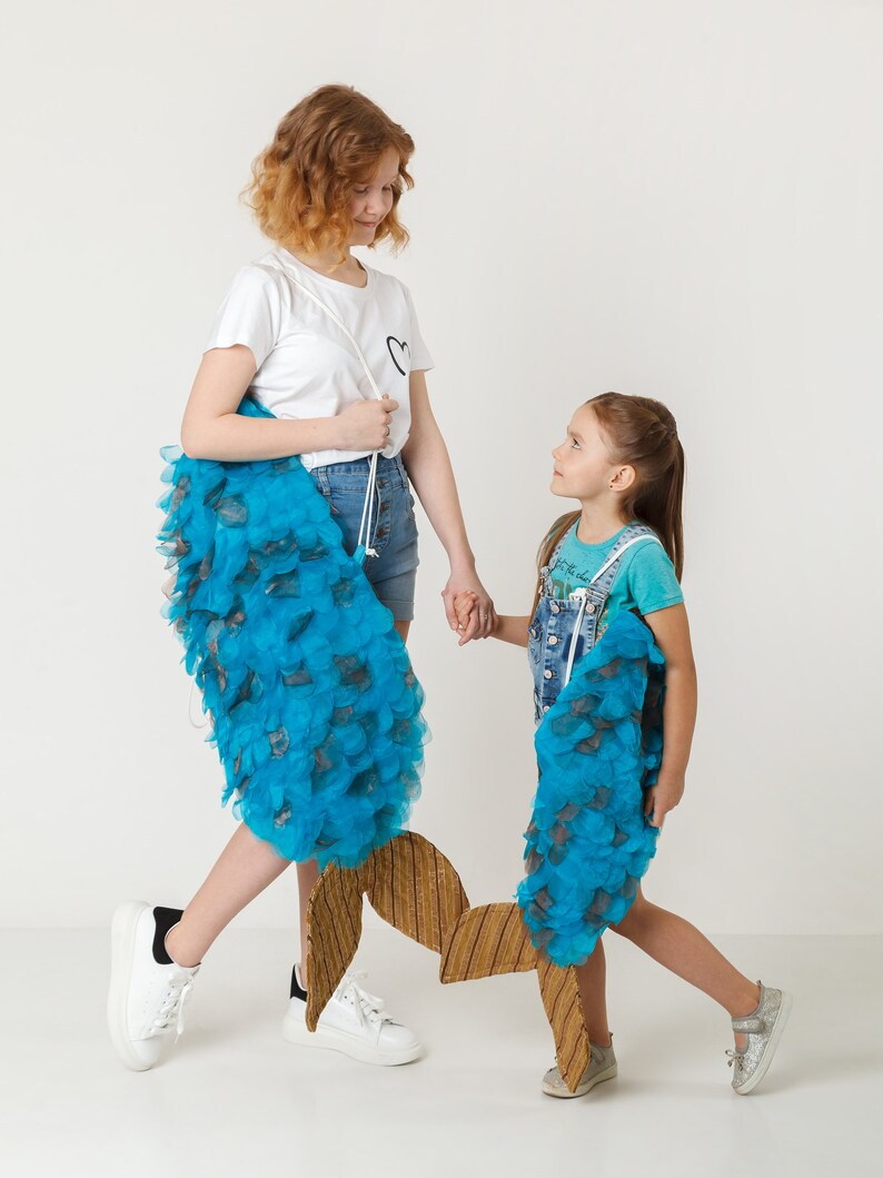 May include: Two girls are holding blue and brown fish-shaped props. The girl on the left is wearing a white t-shirt and denim shorts. The girl on the right is wearing a blue and white striped shirt and denim overalls.