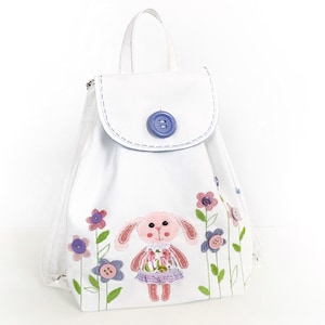 May include: White canvas backpack with a pink bunny and flowers embroidered on the front. The backpack has a blue button closure and a white drawstring.