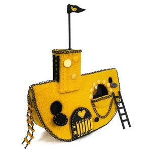 Pode incluir: Um submarino amarelo de feltro com guarnições pretas e uma pequena bandeira preta. O submarino tem uma pequena porta com uma janela em forma de coração e uma escada lateral.