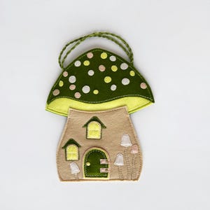 May include: A whimsical felt mushroom house ornament. The house features a tan body with a green door and windows, topped with a green mushroom cap dotted with white, pink, and yellow circles. A green braided loop is attached for hanging.