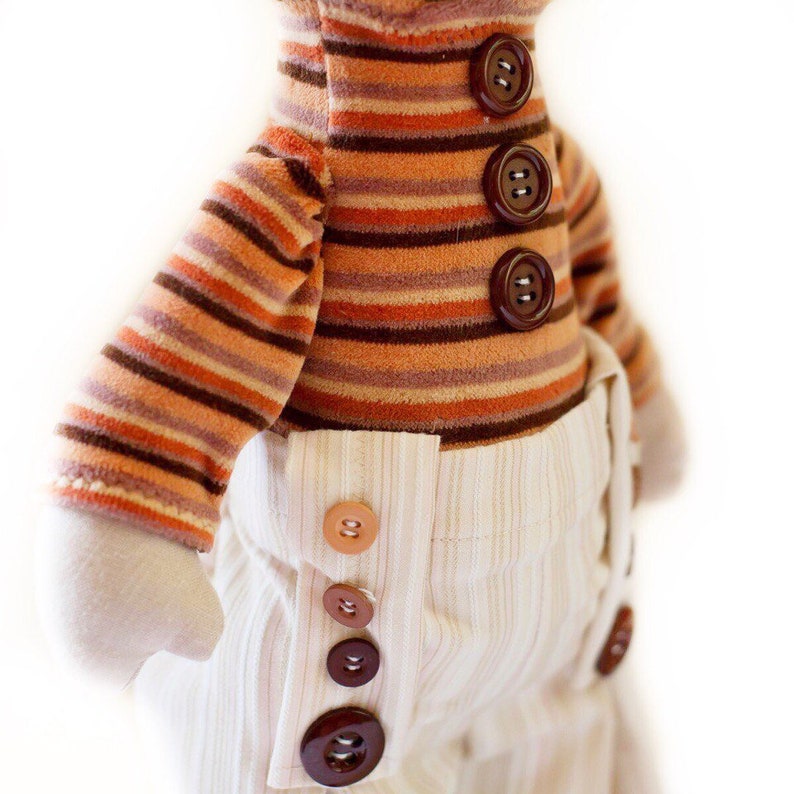 May include: A handmade doll with a striped orange and brown shirt and white pants with brown buttons. The doll has a simple, childlike design.