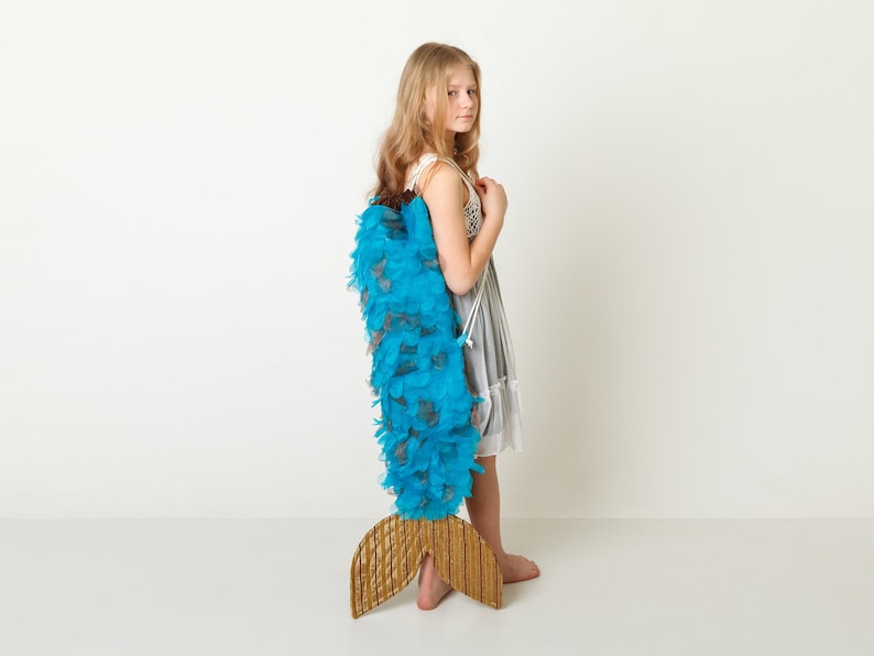 May include: A young girl wearing a white dress with a blue and brown feathered mermaid tail.