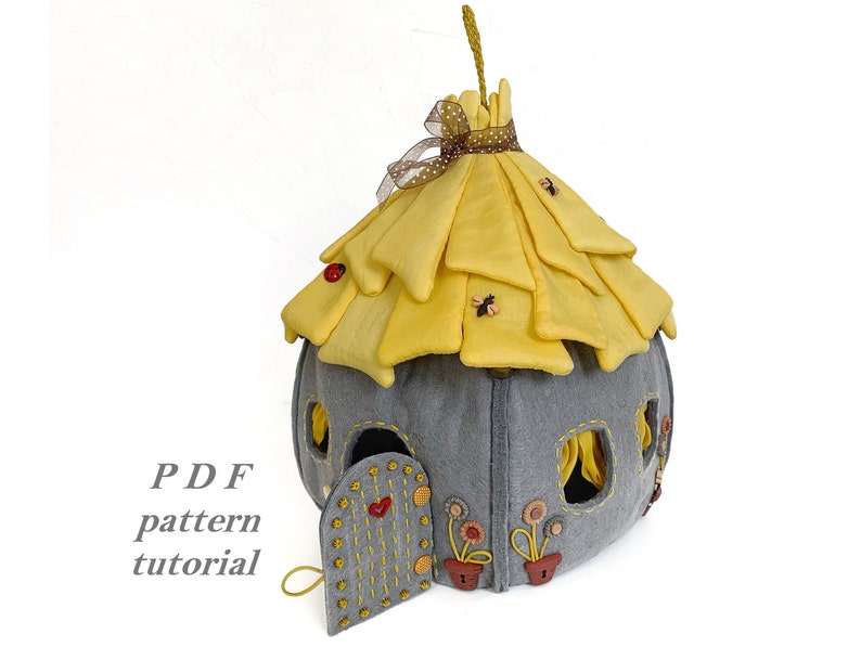 May include: A felt house with a yellow thatched roof, gray walls, and a small door. The house has windows and is decorated with flowers and bees. The text "PDF pattern tutorial" is visible in the image.
