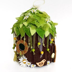 K&ouml;nnte beinhalten: Ein braunes Filz-Feenhaus mit einem gr&uuml;nen Filzdach, verziert mit wei&szlig;en Bl&uuml;ten, gr&uuml;nen Bl&auml;ttern und gelben und gr&uuml;nen Perlen. Das Haus hat ein kleines rundes Fenster und eine Holzvogel-Ausschneidung.