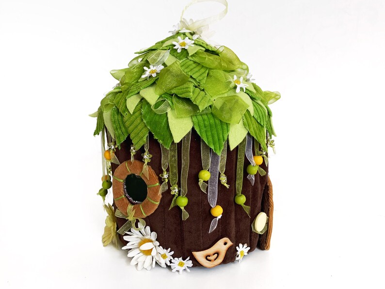 K&ouml;nnte beinhalten: Ein braunes Filz-Feenhaus mit einem gr&uuml;nen Filzdach, verziert mit wei&szlig;en Bl&uuml;ten, gr&uuml;nen Bl&auml;ttern und gelben und gr&uuml;nen Perlen. Das Haus hat ein kleines rundes Fenster und eine Holzvogel-Ausschneidung.