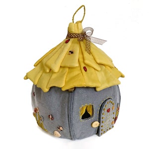 May include: A gray felt fairy house with a yellow felt roof, decorated with buttons, beads, and embroidered details. The roof has a brown ribbon tied around it.