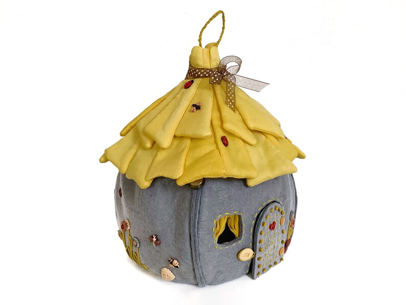 May include: A gray felt fairy house with a yellow felt roof, decorated with buttons, beads, and embroidered details. The roof has a brown ribbon tied around it.