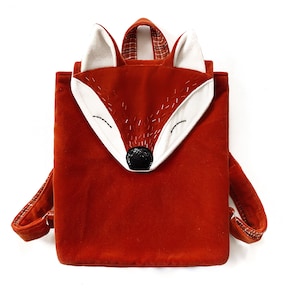 Può includere: Uno zaino rosso a forma di volpe con orecchie bianche e naso nero. Lo zaino è fatto di un tessuto morbido e peluche.