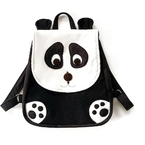 May include: A black and white panda-themed backpack with a plush face and paw prints. The backpack has a white face with black eyes and a brown nose. The backpack has black straps and a black body with white paw prints.