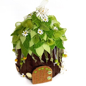 K&ouml;nnte beinhalten: Ein braunes Filz-Feenhaus mit einem gr&uuml;nen Filzdach, verziert mit wei&szlig;en Filzblumen und gr&uuml;nen Filzbl&auml;ttern. Das Haus hat eine kleine braune Filzt&uuml;r mit einem gr&uuml;nen Filzrahmen.