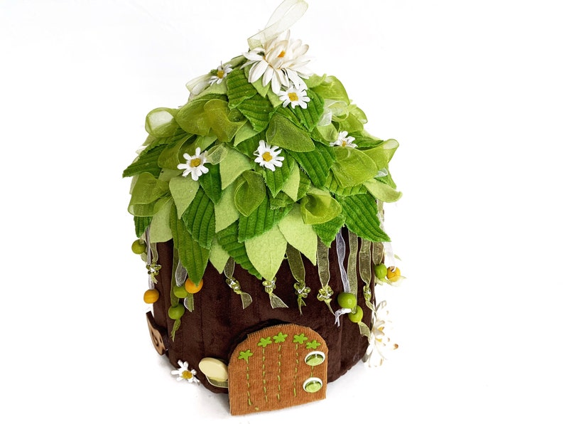 K&ouml;nnte beinhalten: Ein braunes Filz-Feenhaus mit einem gr&uuml;nen Filzdach, verziert mit wei&szlig;en Filzblumen und gr&uuml;nen Filzbl&auml;ttern. Das Haus hat eine kleine braune Filzt&uuml;r mit einem gr&uuml;nen Filzrahmen.