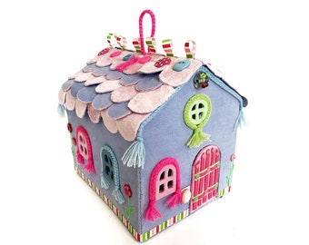 May include: A blue felt fairy house with a pink felt roof, pink and blue windows, a red door, and green felt accents. The house has a loop at the top for hanging.