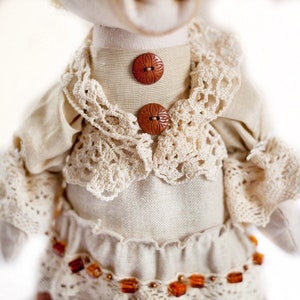 May include: A handmade doll wearing a white linen dress with crocheted lace trim and brown buttons. The doll has a white crocheted collar and a brown beaded belt.