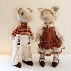May include: Two handmade pig dolls, one wearing a striped shirt and overalls, the other wearing a brown dress with lace trim.